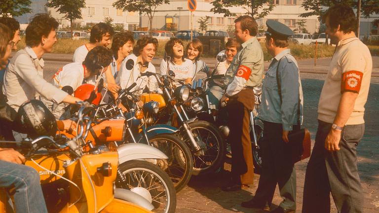 "Jugend in der DDR - Vom Stillstand zum Widerstand": Mehrere Jugendliche sitzen an einer Straße auf ihren Mopeds und sprechen mit drei DDR-Volkspolizisten.