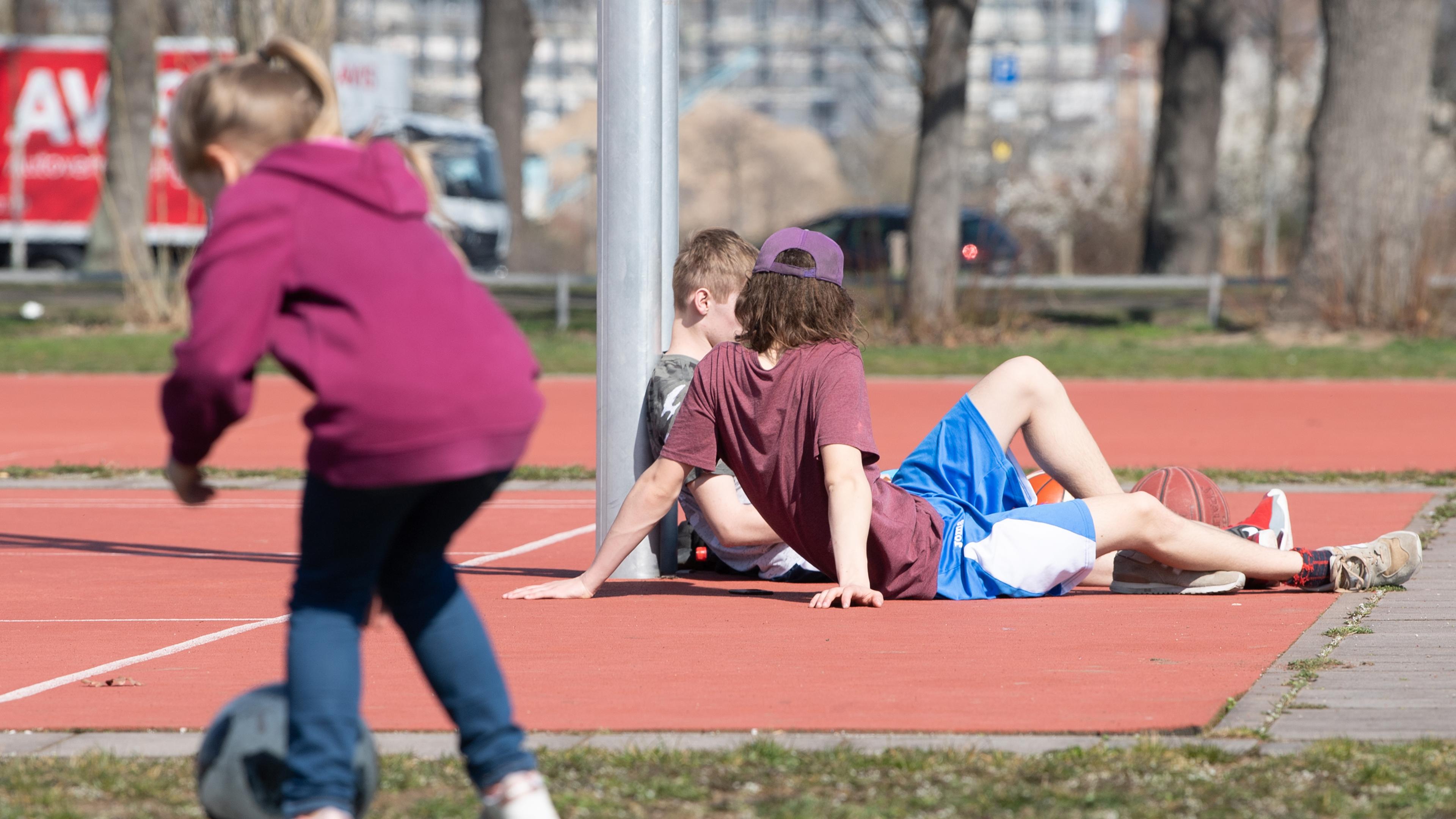 Symbolbild: Ein Kind und zwei Jugendliche stehen und sitzen auf einem Sportplatz.  