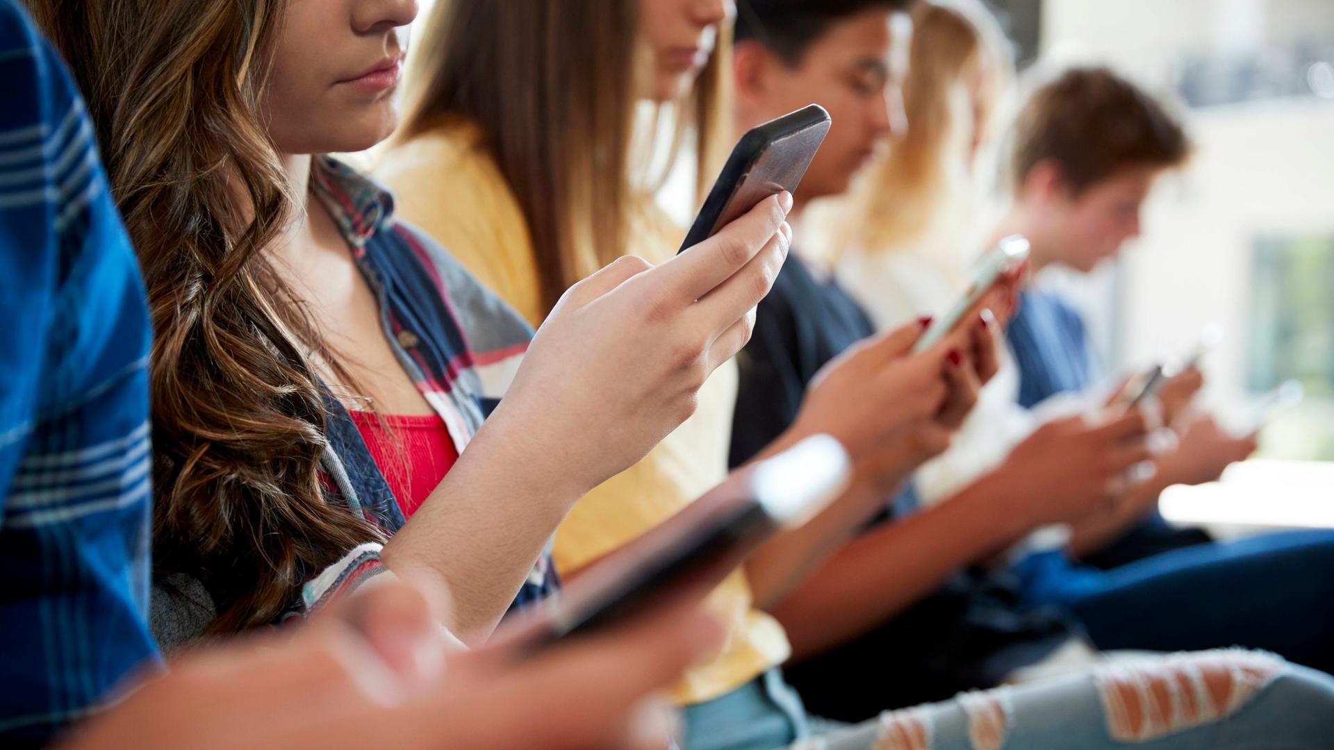 Eine Gruppe Jugendlicher sitzt nebeneinander und schaut auf Smartphone.