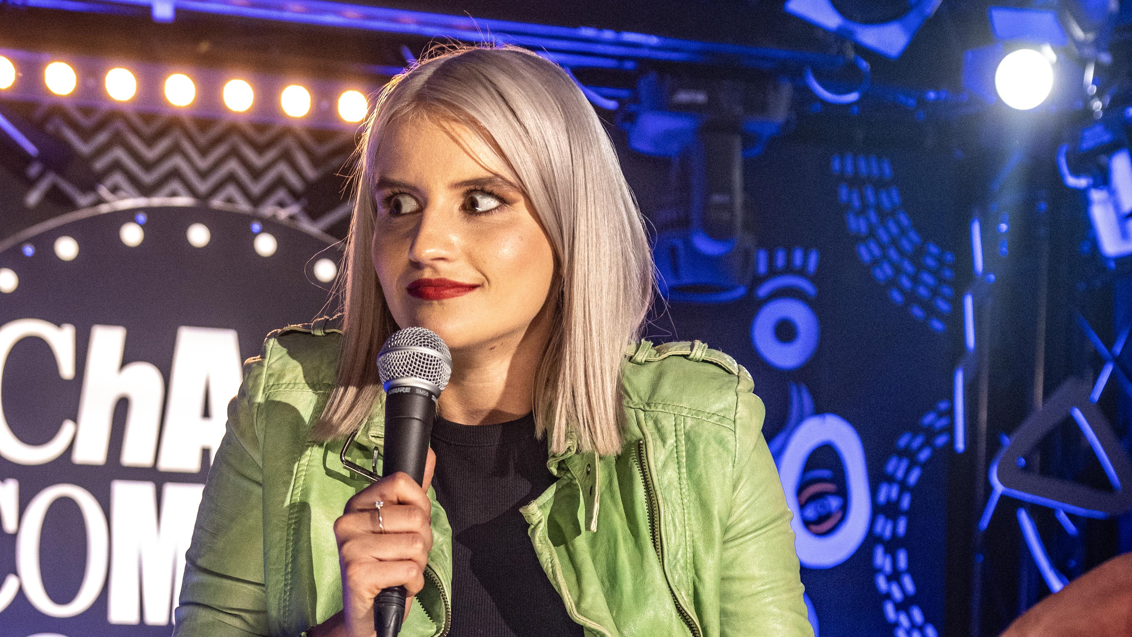 Julia Brandner steht auf der Bühne und zieht die Schultern nach oben. Sie trägt eine grüne Jacke.