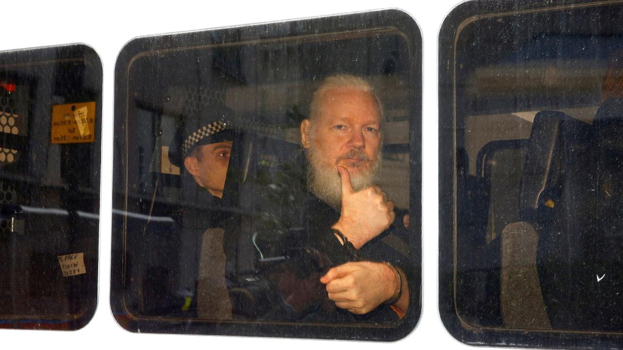 Julian Assange sitzt in einem Polizeiwagen. Er schaut aus dem Fenster des Autos und zeigt einen Daumen nach oben.