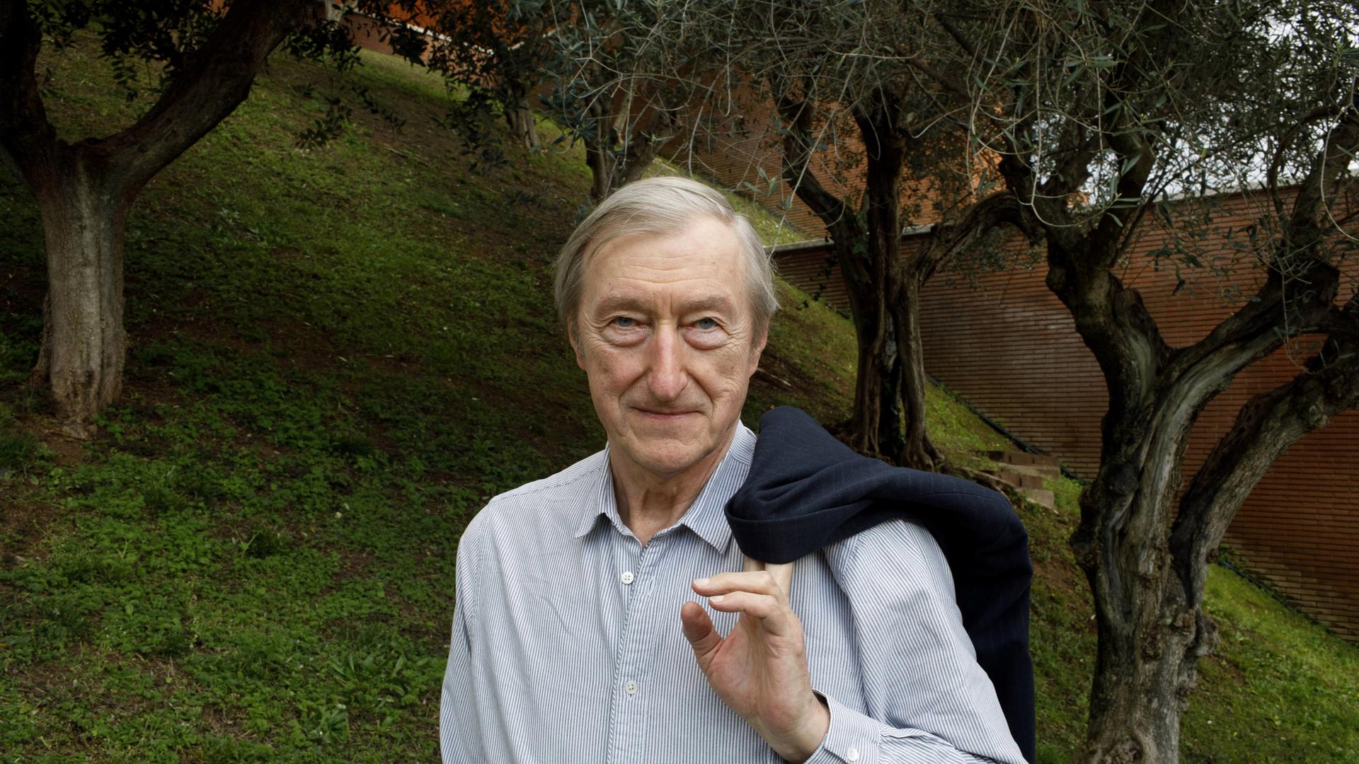 In der Mitte des Bildes steht ein alter Mann in einem gestreiften Hemd. Er blickt leicht schmunzelnd frontal in die Richtung des Betrachters und hat seinen Mantel über der Schulter hängen und hält ihn mit einer Hand fest. Im Hintergrund ist eine Wiese mit Bäumen und rechts eine Mauer zu erkennen.