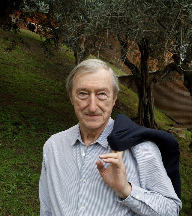 In der Mitte des Bildes steht ein alter Mann in einem gestreiften Hemd. Er blickt leicht schmunzelnd frontal in die Richtung des Betrachters und hat seinen Mantel über der Schulter hängen und hält ihn mit einer Hand fest. Im Hintergrund ist eine Wiese mit Bäumen und rechts eine Mauer zu erkennen.