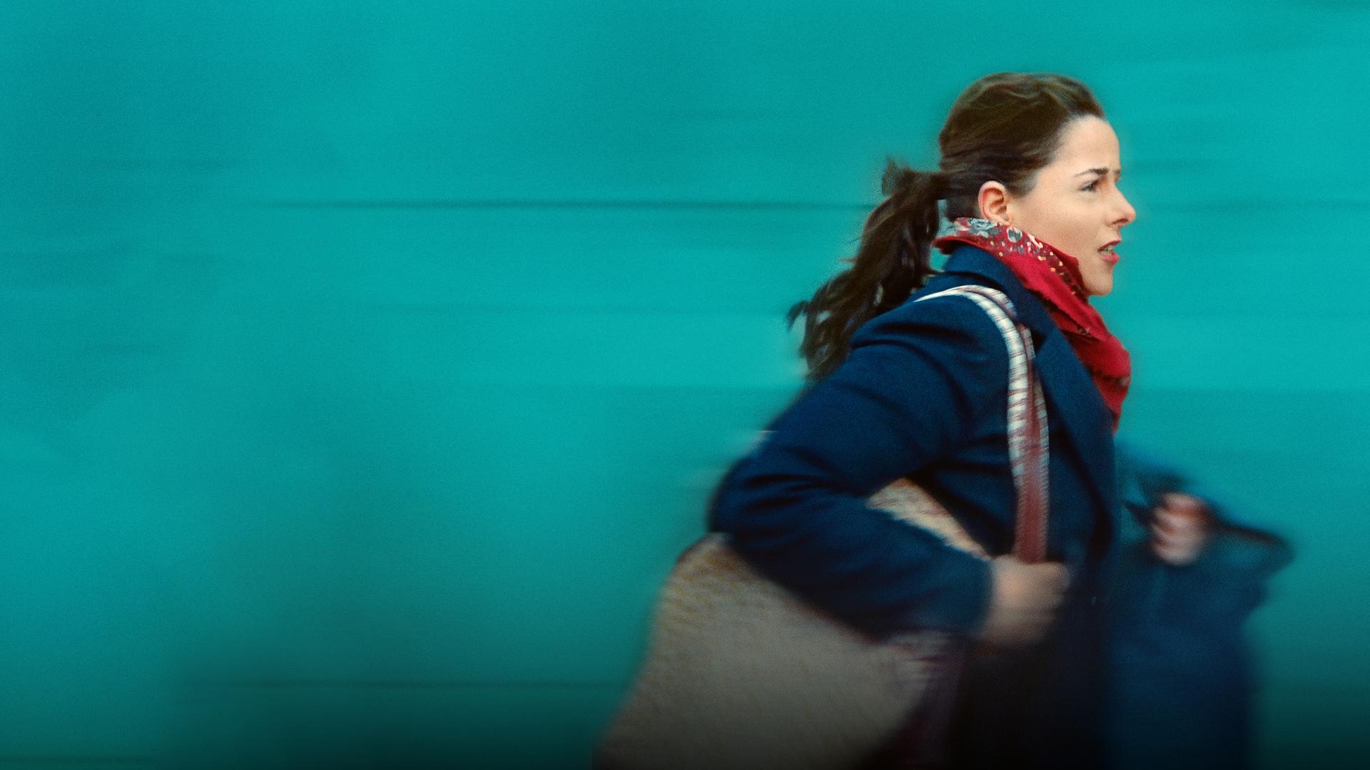 Nahaufnahme: Julie, bekeidet mit einem dunkelblauen Manten, roten Halstuch und einer Tasche über der Schulter, rennt. Sie schaut besorgt, fast verzweifelt.