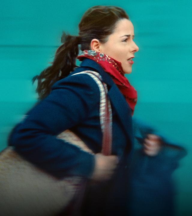 Nahaufnahme: Julie, bekeidet mit einem dunkelblauen Manten, roten Halstuch und einer Tasche über der Schulter, rennt. Sie schaut besorgt, fast verzweifelt.