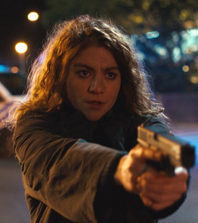 "Juliet - Die Tote im Kanal": Polizistin Juliet Dumon (Charlotte De Bruyne) steht im Dunkeln vor einem Gebäude und einem Auto. Sie hält ihre Pistole in beiden Händen und richtet sie zur Seite.