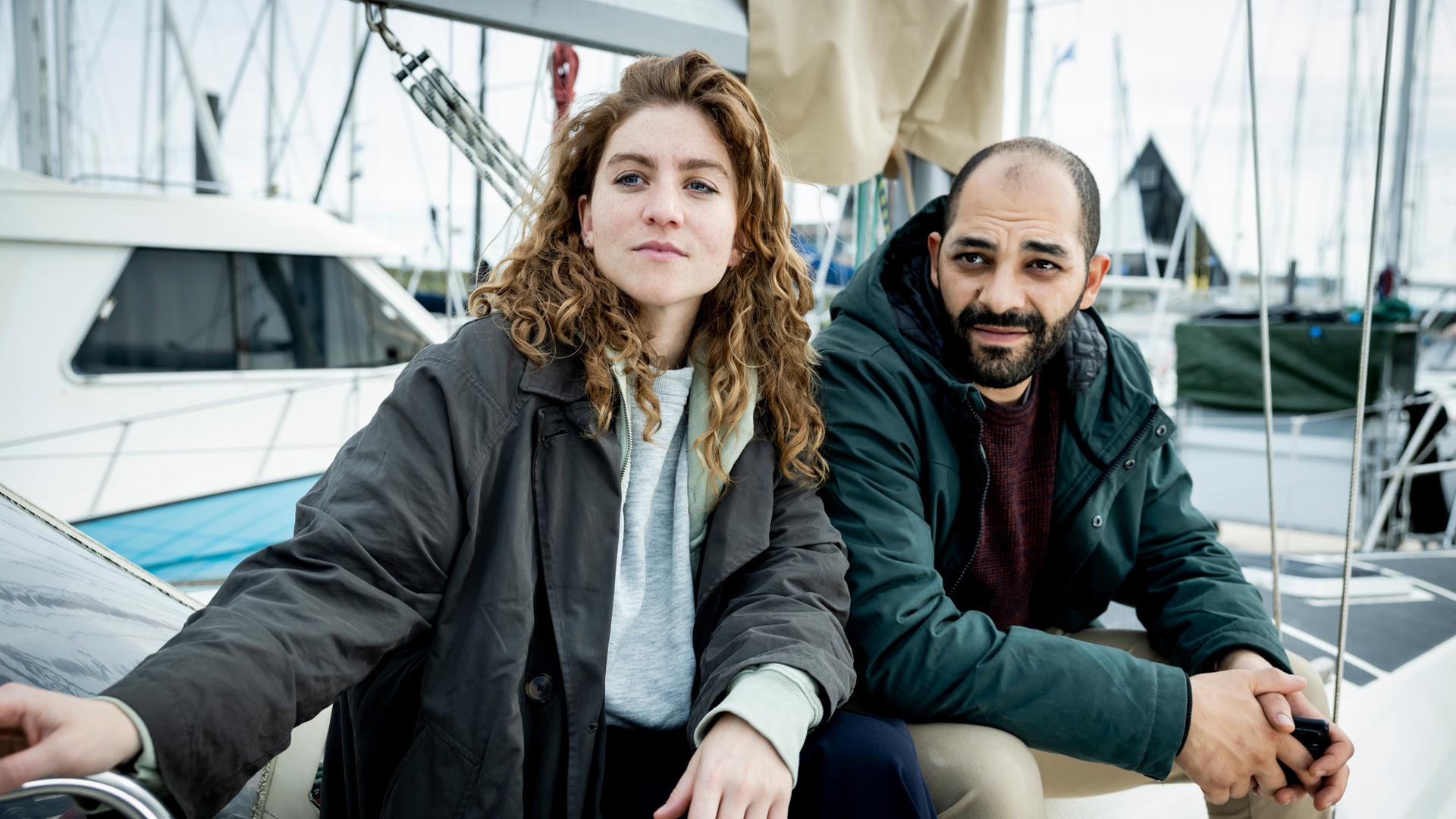 "Juliet - Kaltes Herz": Die Ermittler Juliet Dumon (Charlotte De Bruyne) und Jamal Haout (Nabil Mallat) sitzen nebeneinander am Hafen. Im Hintergrund liegen mehrere Segelboote.