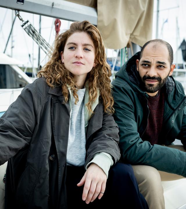 "Juliet - Kaltes Herz": Die Ermittler Juliet Dumon (Charlotte De Bruyne) und Jamal Haout (Nabil Mallat) sitzen nebeneinander am Hafen. Im Hintergrund liegen mehrere Segelboote.