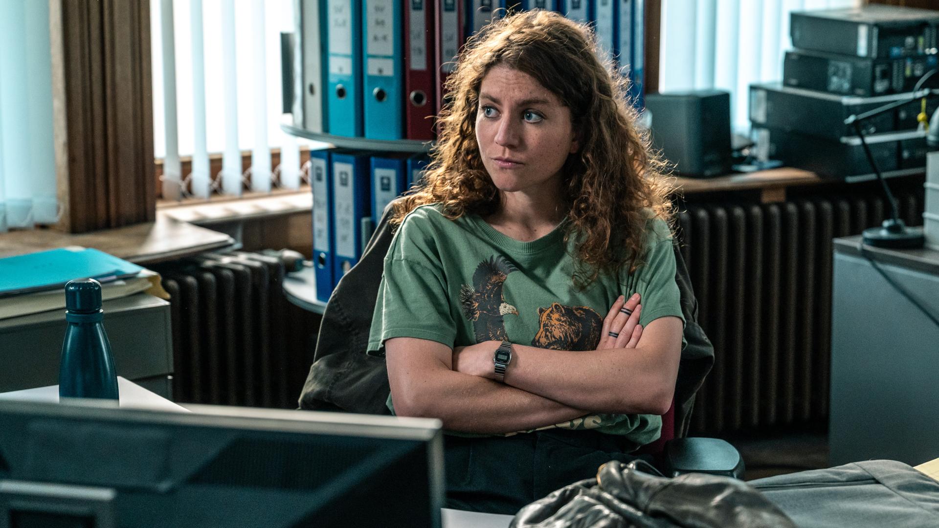 "Juliet - Roter Himmel": Im Büro: Juliet (Charlotte De Bruyne) sitzt in einem dunkelgrünen T-Shirt, das mit einem Adler- und einem Bären-Motiv bedruckt ist, an ihrem Schreibtisch und sinniert über ihren Fall nach.