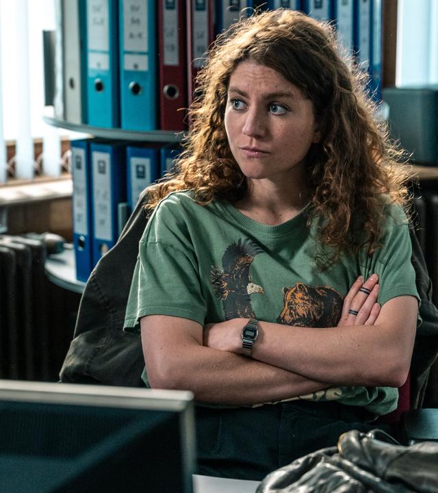 "Juliet - Roter Himmel": Im Büro: Juliet (Charlotte De Bruyne) sitzt in einem dunkelgrünen T-Shirt, das mit einem Adler- und einem Bären-Motiv bedruckt ist, an ihrem Schreibtisch und sinniert über ihren Fall nach.