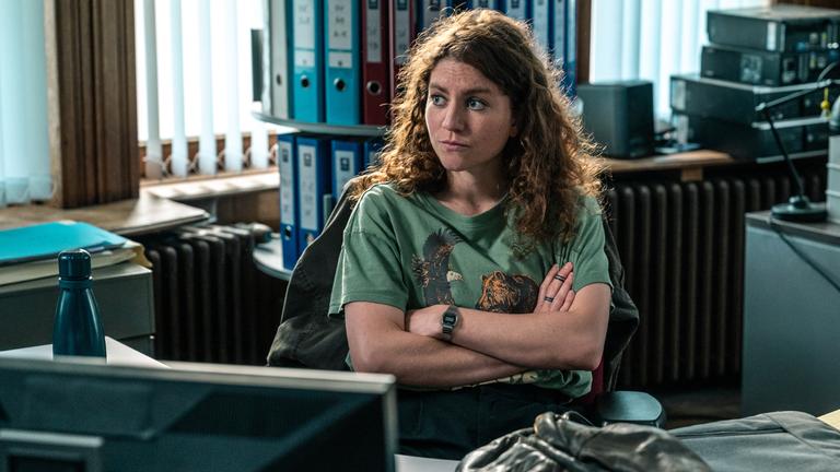 "Juliet - Roter Himmel": Im Büro: Juliet (Charlotte De Bruyne) sitzt in einem dunkelgrünen T-Shirt, das mit einem Adler- und einem Bären-Motiv bedruckt ist, an ihrem Schreibtisch und sinniert über ihren Fall nach.