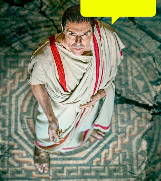 Spielszene, Blick von oben: Gaius Julius Cäsar trägt eine helle römische Toga mit roten Streifen. Der gemusterte Boden, auf dem er steht, bricht seitlich weg.