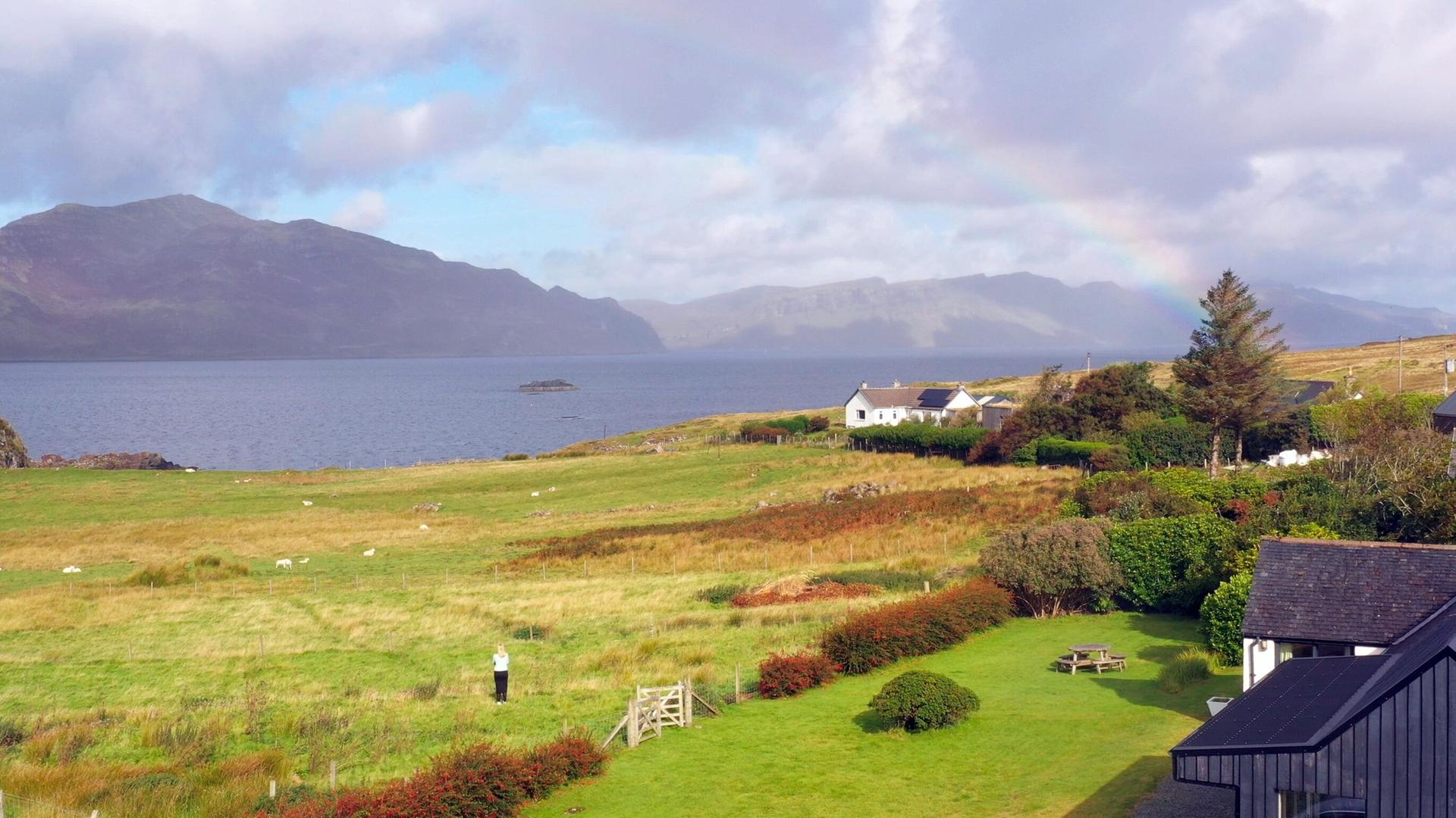Das Bild zeigt eine weitläufige Landschaft auf der schottischen Insel Raasay. Im Vordergrund ist eine grüne Wiese mit einigen niedrigen, buschigen Pflanzen sichtbar. Eine Person steht auf der Wiese, die in hellen Kleidungsstücken gekleidet ist. Zum Hintergrund hin erstreckt sich sanft das Land bis zu einem Gewässer, das ruhig ist und im Licht schimmert.  Auf der anderen Seite des Wassers sind bewaldete und hügelige Berge zu sehen, die den Horizont schmücken. In der Nähe des Wassers steht ein weißes, einstöckiges Haus, das von Bäumen und Sträuchern umgeben ist. Ein kleiner Radweg oder Zaun aus Holz befindet sich in der unteren Bildmitte, der das Bild in verschiedene Bereiche unterteilt.  Am Himmel sind einige Wolken zu sehen, die teilweise von Sonnenstrahlen durchbrochen werden. Ein schwacher Regenbogen ist sichtbar, was dem Bild eine idyllische und friedliche Atmosphäre verleiht. Die Gesamtkomposition vermittelt ein Gefühl von Einsamkeit und Abgeschiedenheit in einer malerischen Umgebung.