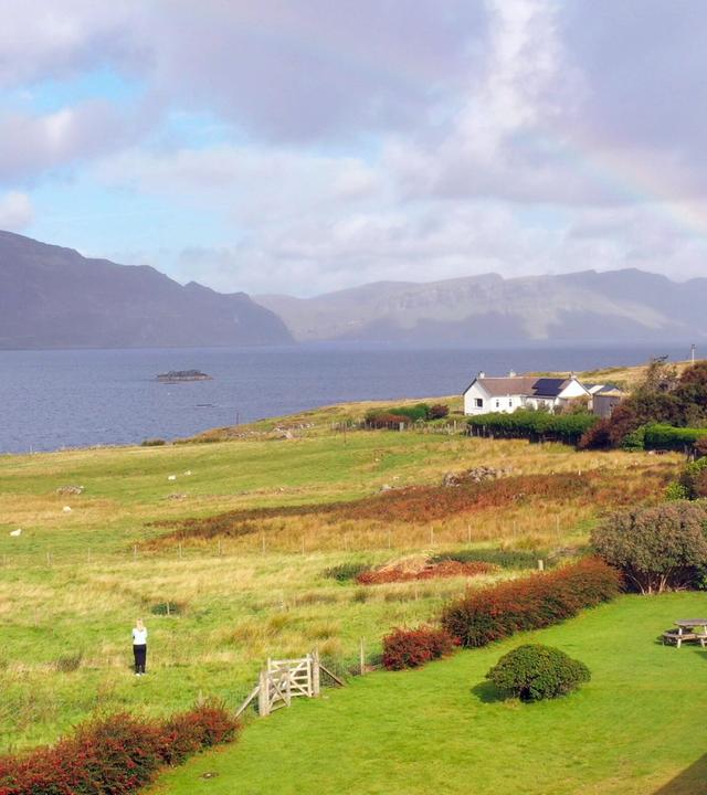 Das Bild zeigt eine weitläufige Landschaft auf der schottischen Insel Raasay. Im Vordergrund ist eine grüne Wiese mit einigen niedrigen, buschigen Pflanzen sichtbar. Eine Person steht auf der Wiese, die in hellen Kleidungsstücken gekleidet ist. Zum Hintergrund hin erstreckt sich sanft das Land bis zu einem Gewässer, das ruhig ist und im Licht schimmert.  Auf der anderen Seite des Wassers sind bewaldete und hügelige Berge zu sehen, die den Horizont schmücken. In der Nähe des Wassers steht ein weißes, einstöckiges Haus, das von Bäumen und Sträuchern umgeben ist. Ein kleiner Radweg oder Zaun aus Holz befindet sich in der unteren Bildmitte, der das Bild in verschiedene Bereiche unterteilt.  Am Himmel sind einige Wolken zu sehen, die teilweise von Sonnenstrahlen durchbrochen werden. Ein schwacher Regenbogen ist sichtbar, was dem Bild eine idyllische und friedliche Atmosphäre verleiht. Die Gesamtkomposition vermittelt ein Gefühl von Einsamkeit und Abgeschiedenheit in einer malerischen Umgebung.