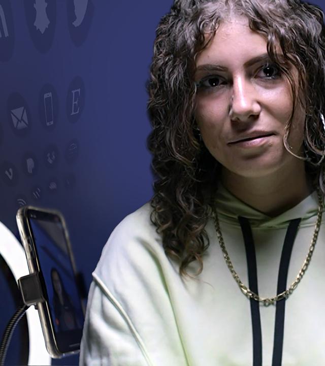 Porträtaufnahme: Laura, eine junge Frau mit Locken und gelbem Pullover, sitzt vor schwarzem Hintergrund und schaut in die Kamera.