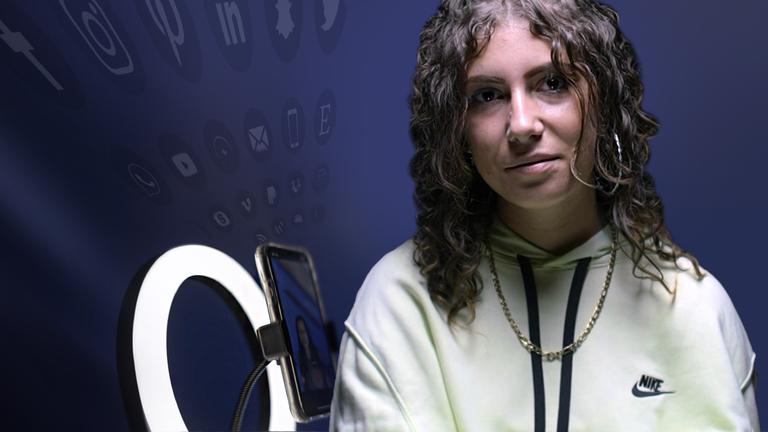 Porträtaufnahme: Laura, eine junge Frau mit Locken und gelbem Pullover, sitzt vor schwarzem Hintergrund und schaut in die Kamera.