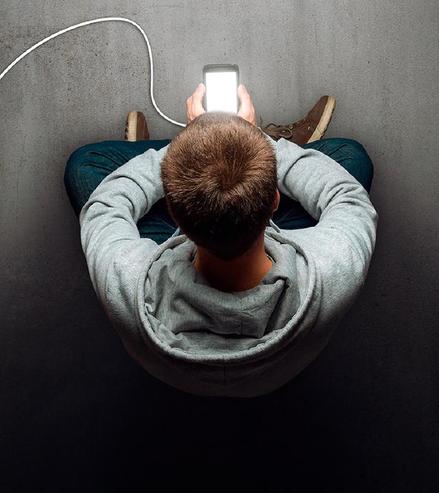 Ein Junge im grauen Kapuzenpullover sitzt im Schneidersitz auf dem Boden und schaut auf ein leuchtendes Mobiltelefon, das er in der Hand hält.
