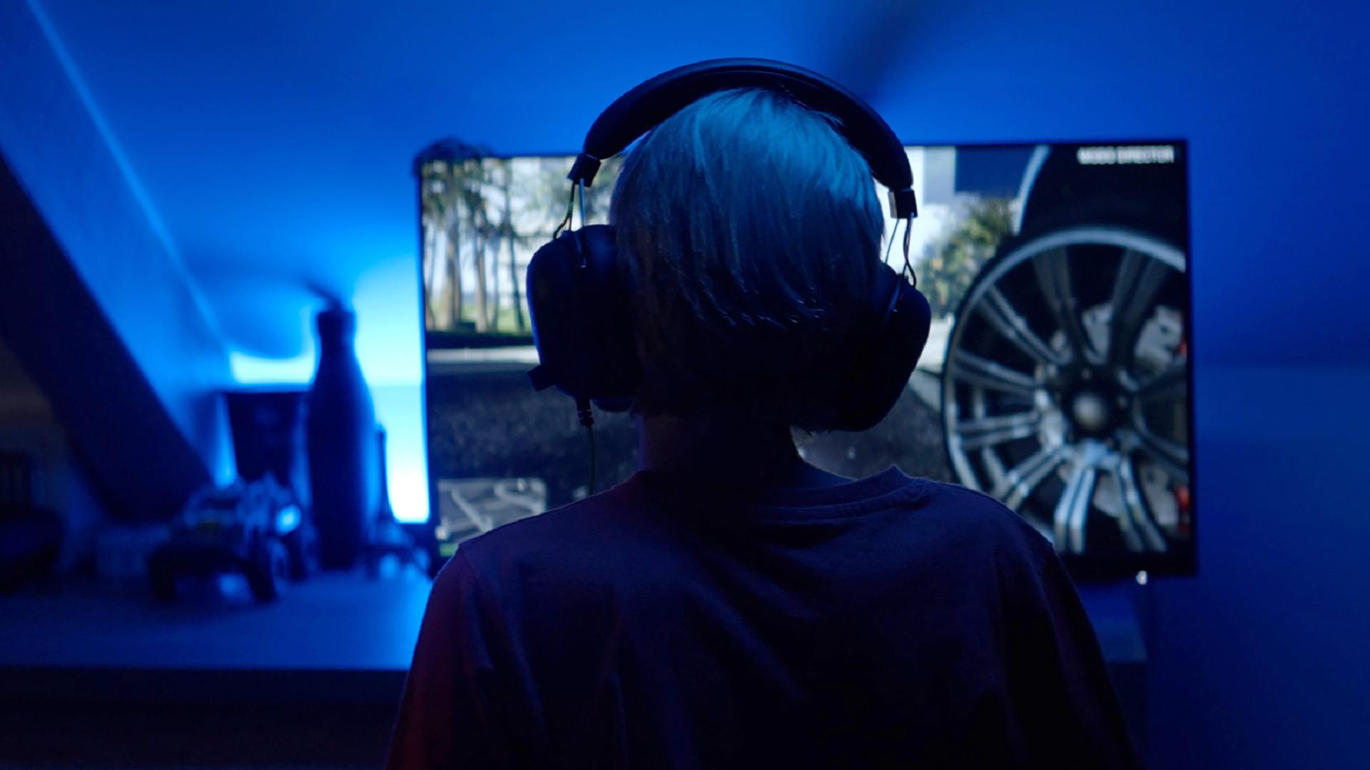 Ein Kind sitzt mit großen Kopfhörern in einem blau beleuchteten Raum vor einem Bildschirm, auf dem Autoreifen zu sehen sind.