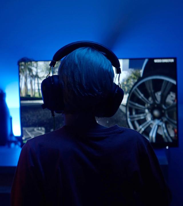Ein Kind sitzt mit großen Kopfhörern in einem blau beleuchteten Raum vor einem Bildschirm, auf dem Autoreifen zu sehen sind.
