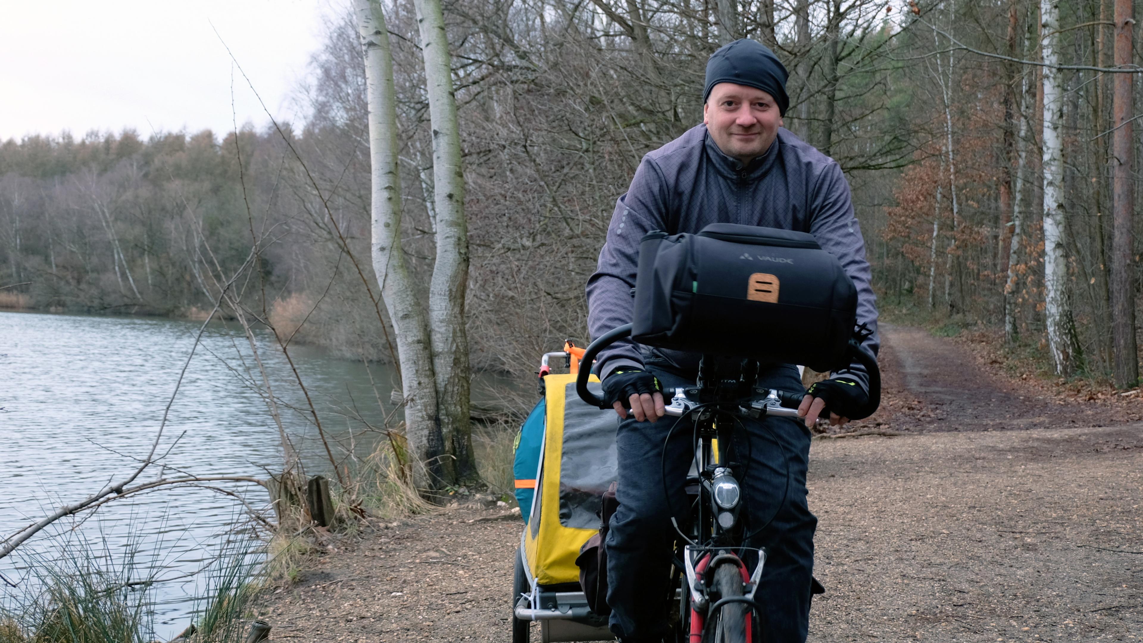Björn ist mittig im Bild auf seinem Fahrrad zu sehen. Er trägt eine schwarze Mütze und eine graue Jacke. Rechts von ihm sieht man das Ufer eines Sees. Links von ihm führt ein Weg in einen Wald.
