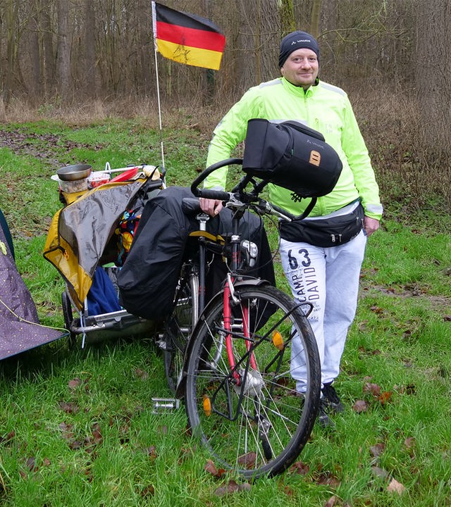 Björn hockt mittig im Bild an einem Camping-Grill. Er trägt eine schwarze Mütze und eine gelbe Jacke. Links von ihm sieht man das Ufer eines Sees. Hinter ihm sieht man sein Fahrrad mit Anhänger und ein Zelt.