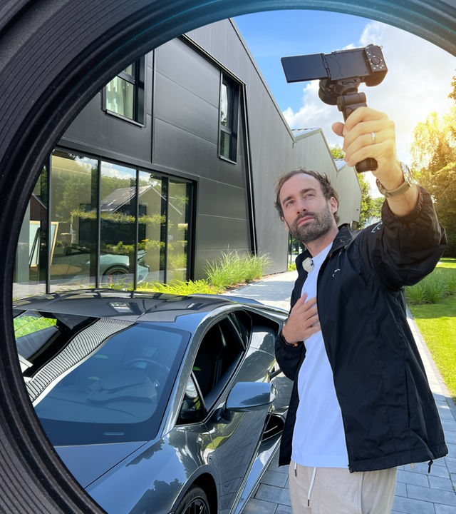 Ein junger Mann hält eine Handykamera hoch und filmt sich mit einem Lamborghini.
