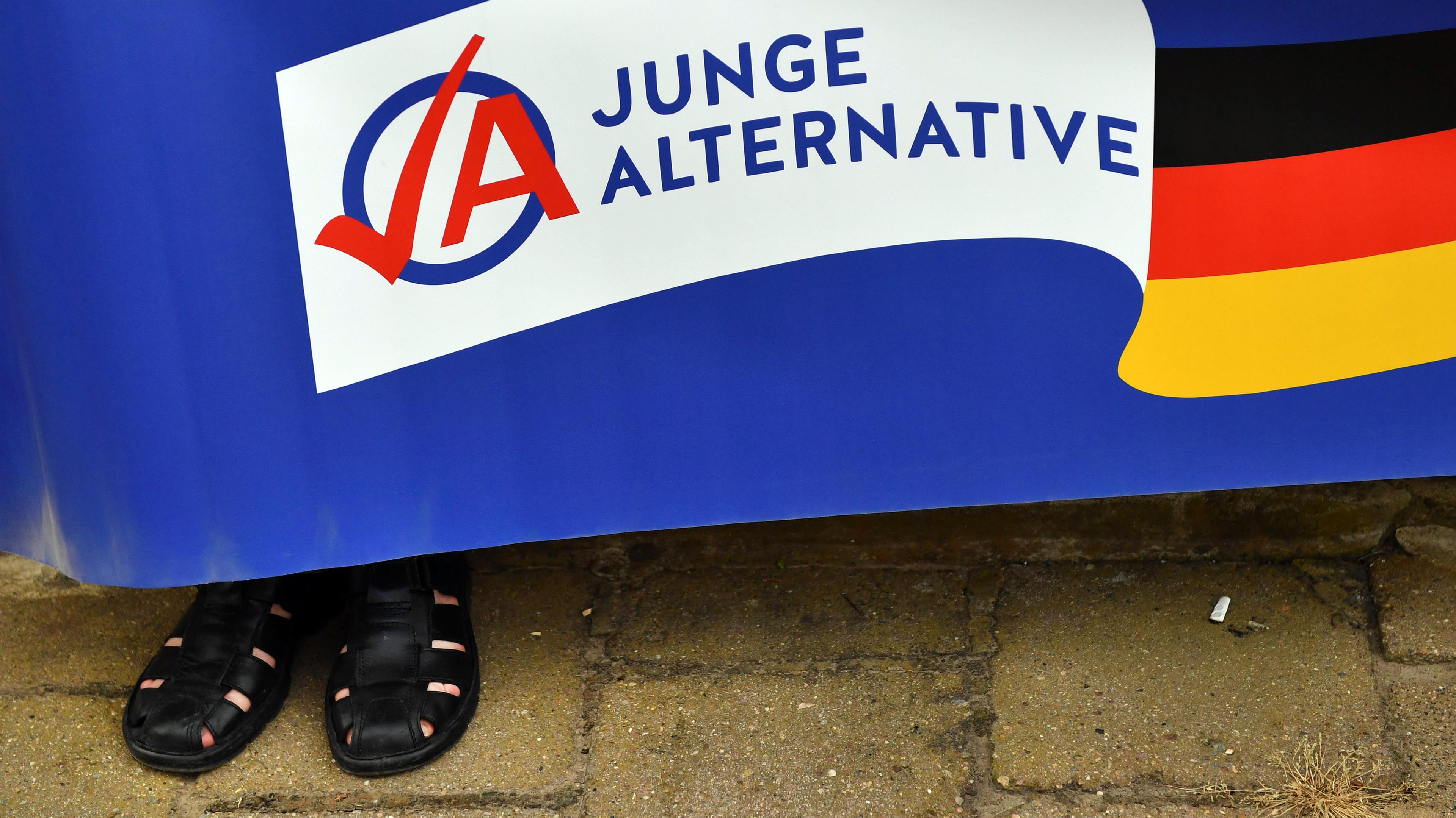 Ein Mann in schwarzen Sandalen hält ein Plakat der AfD-Jugendorganisation Junge Alternative