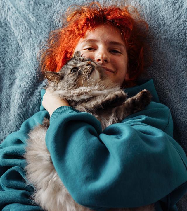 Eine junge Frau mit roten Haaren liegt auf dem Bett und hat eine grau getiegerte Katze im Arm auf sich lieben.