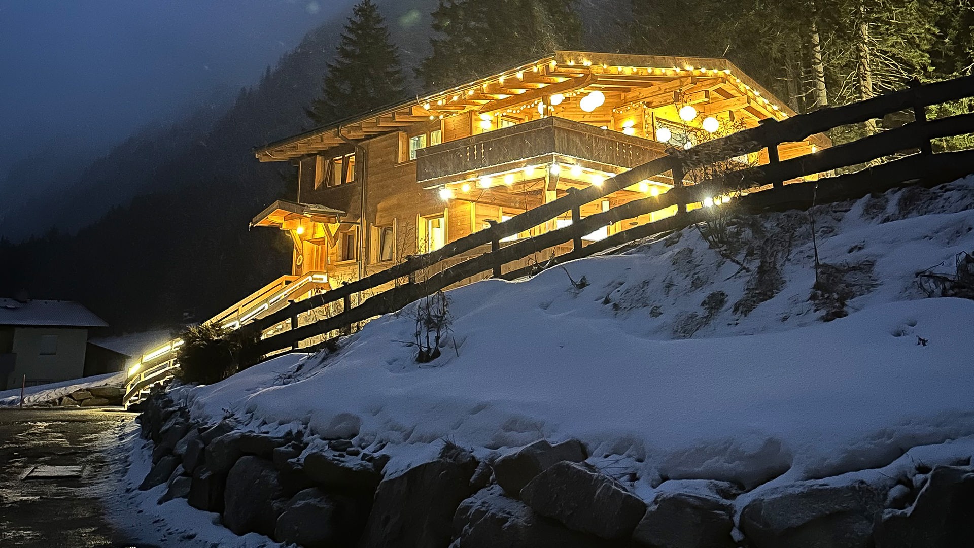 Das im Dunkeln aufgenommene Foto zeigt ein hell erleuchtetes Holz-Chalet mit Lampions und Lichterketten. Eine Straße führt zur Villa, rechts ist ein verschneiter Steinhang mit Holzzaun zu sehen. Im Hintergrund sieht man Tannen und hohe Berge.
