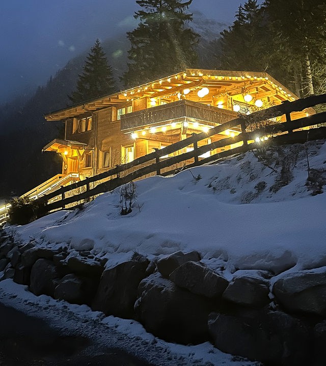 Das im Dunkeln aufgenommene Foto zeigt ein hell erleuchtetes Holz-Chalet mit Lampions und Lichterketten. Eine Straße führt zur Villa, rechts ist ein verschneiter Steinhang mit Holzzaun zu sehen. Im Hintergrund sieht man Tannen und hohe Berge.