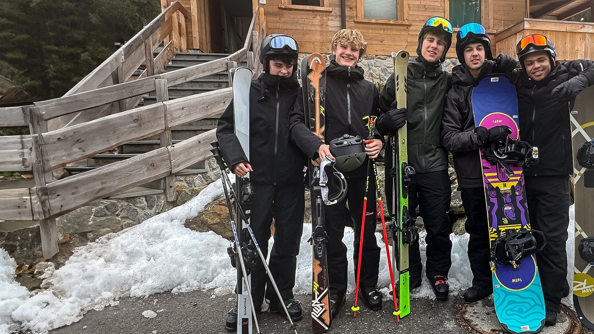 Die fünf Jungs der Schnee-WG Nick, Cay, Luke, Lorian und Azad stehen in Winterkleidung und mit Skiern oder Snowboards in den Händen nebeneinander vor dem Eingang ihres Holz-Chalets. Sie tragen Helme mit Skibrillen und schauen fröhlich in die Kamera. Sie sind offenbar sehr bereit für einen Tag auf der Skipiste.