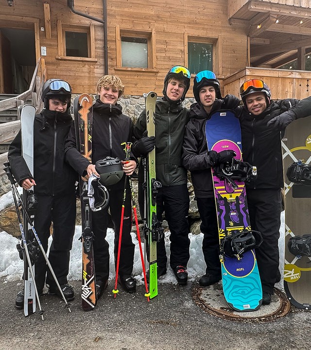 Die fünf Jungs der Schnee-WG Nick, Cay, Luke, Lorian und Azad stehen in Winterkleidung und mit Skiern oder Snowboards in den Händen nebeneinander vor dem Eingang ihres Holz-Chalets. Sie tragen Helme mit Skibrillen und schauen fröhlich in die Kamera. Sie sind offenbar sehr bereit für einen Tag auf der Skipiste.