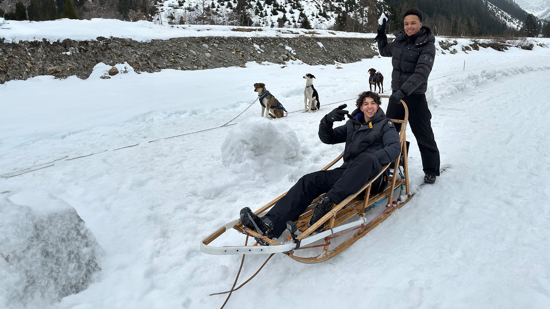 In einer Winterlandschaft sind zwei Jungs in Winterkleidung zu sehen. Lorian sitzt in einem typischen Holzschlitten, der für Schlittenhunde gemacht ist. Allerdings sieht man die Schlittenhunde noch angeleint im Hintergrund im Schnee sitzen. Lorian lacht in die Kamera und macht ein schräges Victory-Zeichen. Hinter ihm auf den Kufen des Schlittens steht Azad und hält sich am Griff des Schlittens fest. Er hat einen Schneeball in der Hand und tut so, als würde er diesen dem Fotografen entgegen werfen.