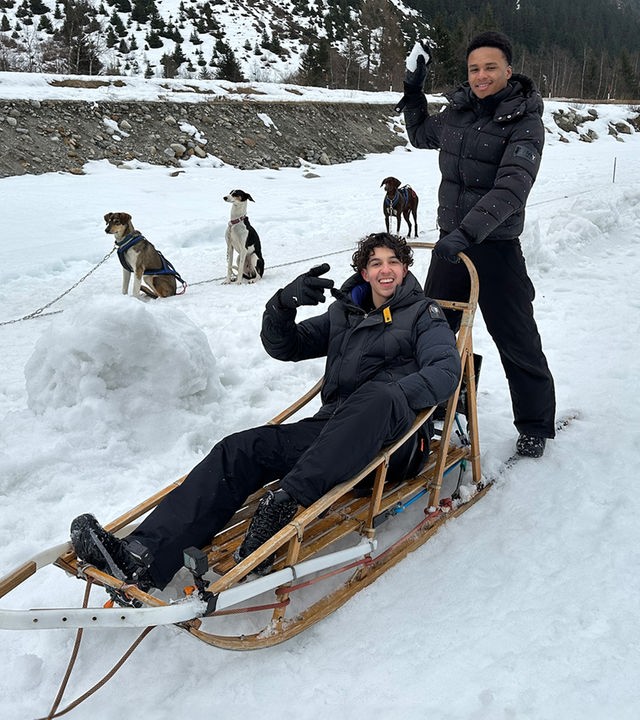 In einer Winterlandschaft sind zwei Jungs in Winterkleidung zu sehen. Lorian sitzt in einem typischen Holzschlitten, der für Schlittenhunde gemacht ist. Allerdings sieht man die Schlittenhunde noch angeleint im Hintergrund im Schnee sitzen. Lorian lacht in die Kamera und macht ein schräges Victory-Zeichen. Hinter ihm auf den Kufen des Schlittens steht Azad und hält sich am Griff des Schlittens fest. Er hat einen Schneeball in der Hand und tut so, als würde er diesen dem Fotografen entgegen werfen.