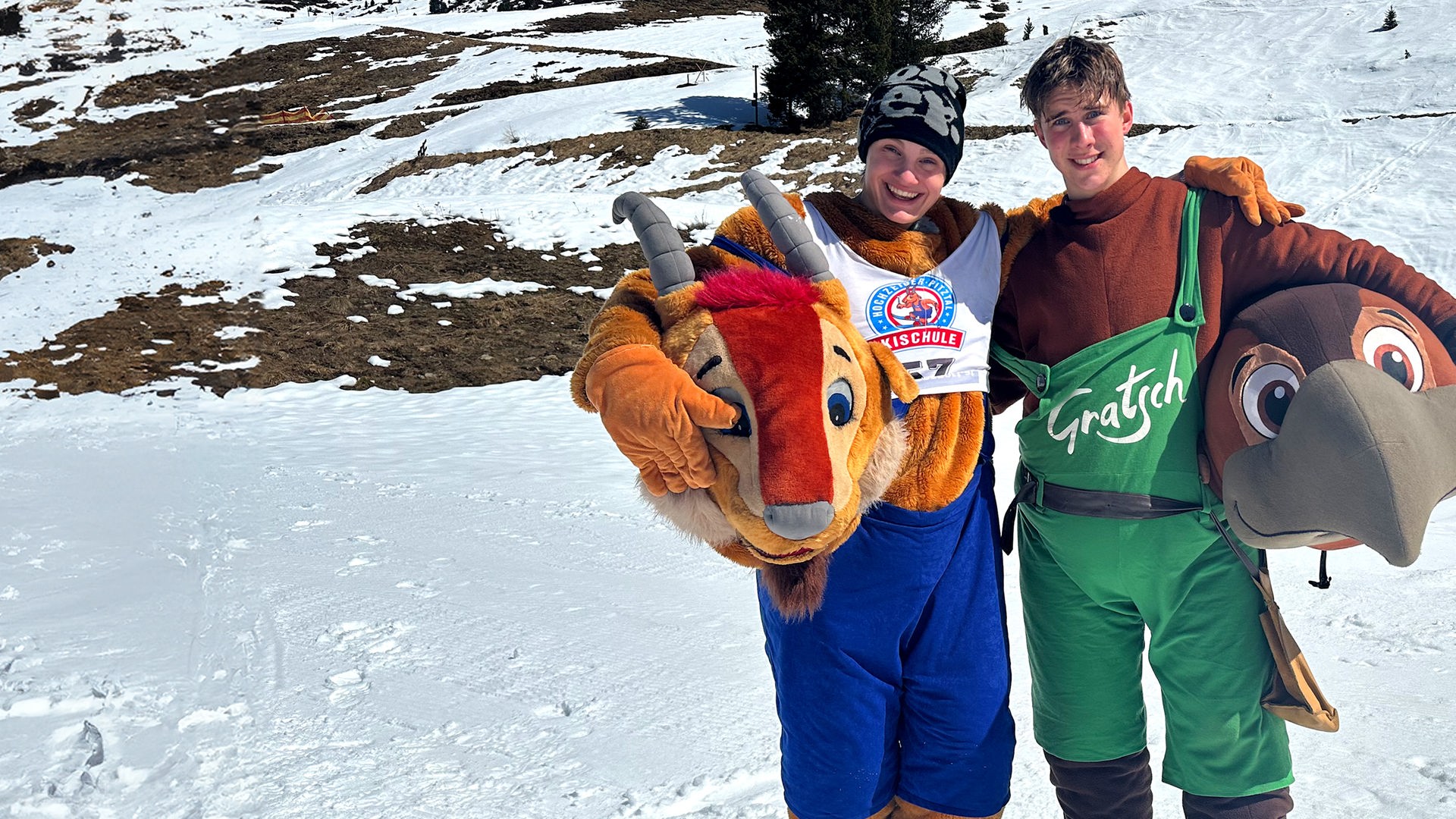 Zwei Jungs der Schnee-WG, Cay und Luke, sind als Maskottchen einer Skischule verkleidet und tragen die übergroßen Köpfe der Tiere in den Händen. Cay trägt einen Ziegenkopf und lacht in die Kamera, Luke einen Vogel. Im Hintergrund ist die Skipiste zu sehen.