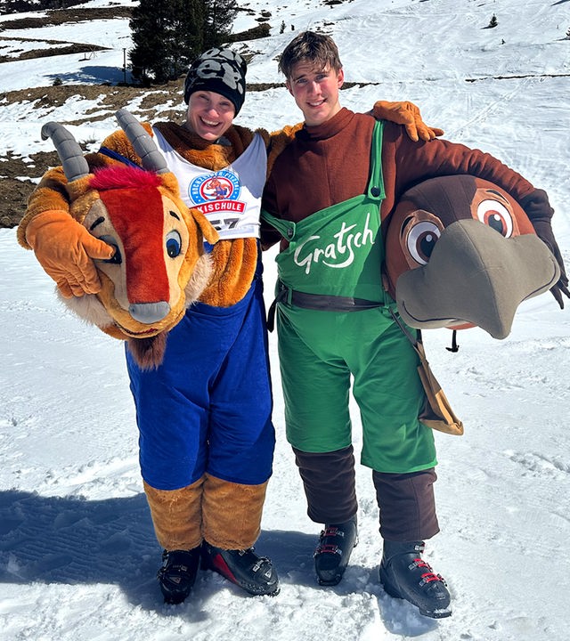 Zwei Jungs der Schnee-WG, Cay und Luke, sind als Maskottchen einer Skischule verkleidet und tragen die übergroßen Köpfe der Tiere in den Händen. Cay trägt einen Ziegenkopf und lacht in die Kamera, Luke einen Vogel. Im Hintergrund ist die Skipiste zu sehen.