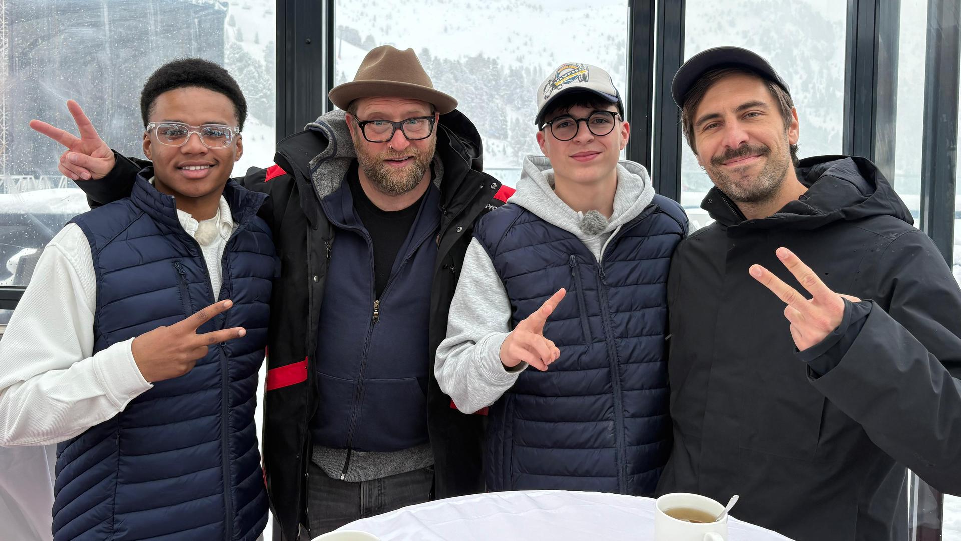 An einem Bistrotisch stehen zwei Jungs mit zwei Männern und grinsen in die Kamera. Alle machen das Victoryzeichen. Es handelt sich um Azad aus der Jungs-WG im Schnee, der neben dem deutschen Musiker Gregor Meyle steht. Daneben steht Nick und sein Idol, der Singer und Songwriter Max Giesinger. Im Hintergrund sieht man durch die großen Fensterscheiben schneebedeckte Berge.