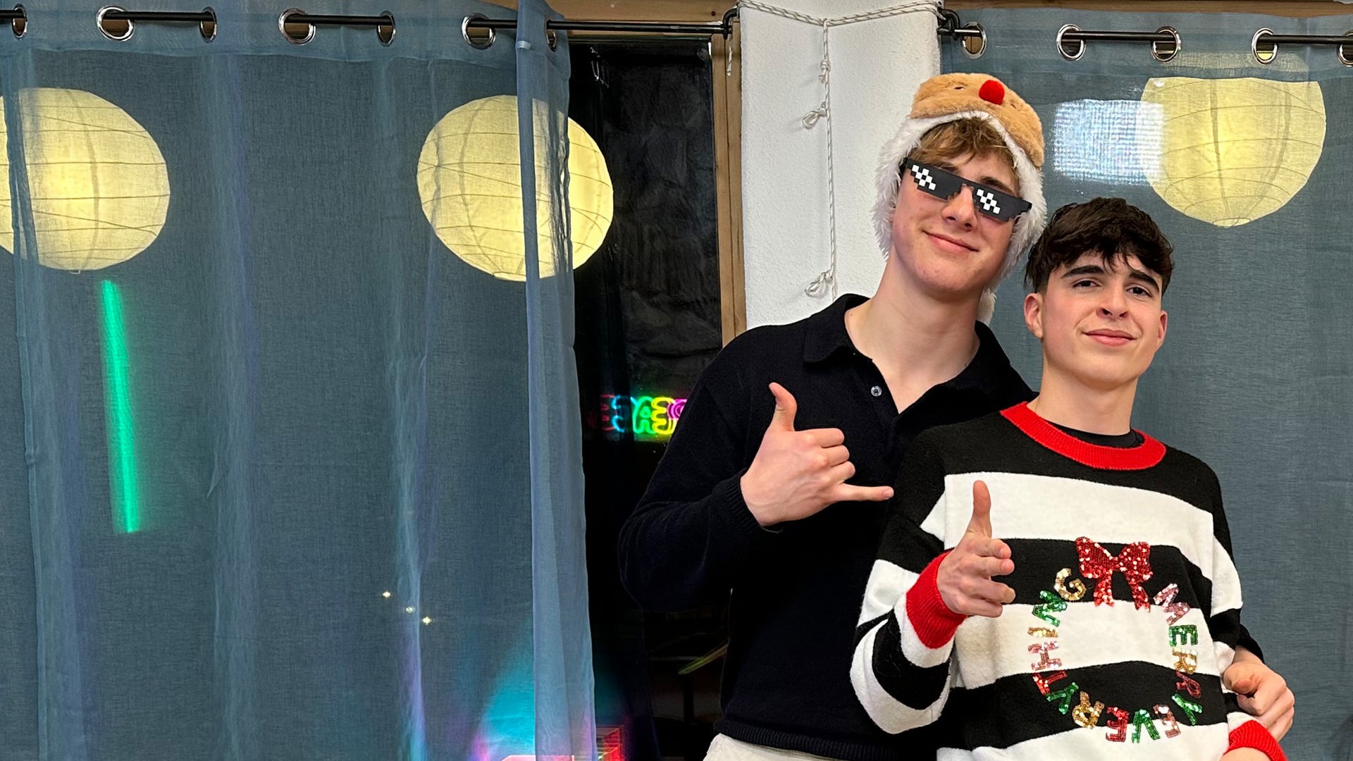 Zwei Jungs stehen aneinander gelehnt vor einer Fensterbank. Im Hintergrund sieht man, dass es draußen bereits dunkel ist und die Lampions vor den Fenstern leuchten. Die Jungs tragen weihnachtliche Outfits. Nick hat einen schwarz-weiß gestreiften Pullover mit rotem Kragen, glitzernder Aufschrift und Schleife an. Luke trägt eine coole schwarze Sonnenbrille mit weißen Punkten und eine wattierte Mütze mit Elchnase.