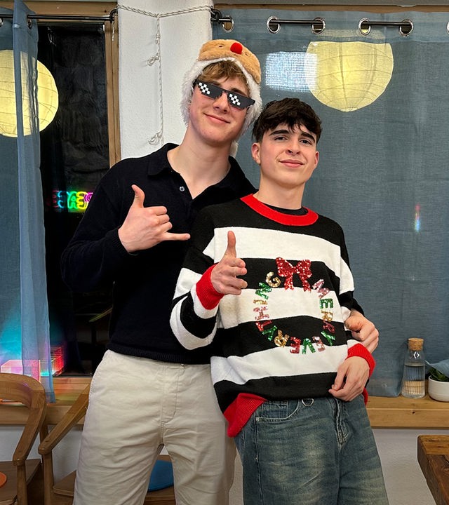 Zwei Jungs stehen aneinander gelehnt vor einer Fensterbank. Im Hintergrund sieht man, dass es draußen bereits dunkel ist und die Lampions vor den Fenstern leuchten. Die Jungs tragen weihnachtliche Outfits. Nick hat einen schwarz-weiß gestreiften Pullover mit rotem Kragen, glitzernder Aufschrift und Schleife an. Luke trägt eine coole schwarze Sonnenbrille mit weißen Punkten und eine wattierte Mütze mit Elchnase.