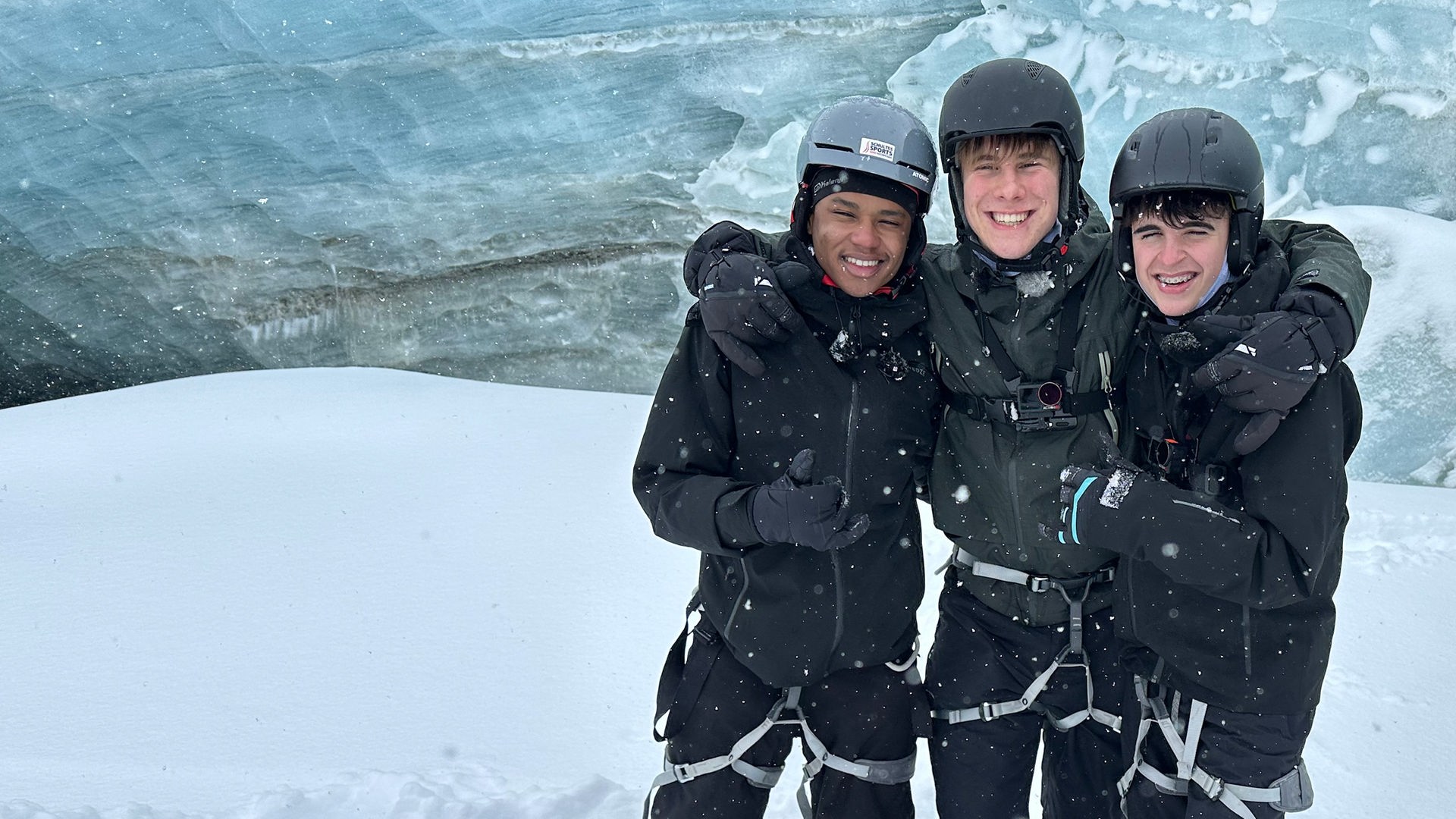 Drei Jungs der Schnee-WG stehen nebeneinander in einer Schneelandschaft. Sie tragen Klettergurte über ihren Skihosen und Helme auf dem Kopf. Azad, Luke und Nick haben die Arme umeinander gelegt und lachen in die Kamera. Hinter ihnen ist die Eiswand einer Gletscherhöhle zu sehen.