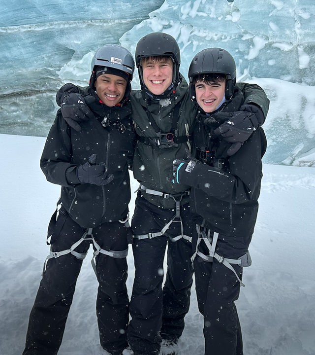 Drei Jungs der Schnee-WG stehen nebeneinander in einer Schneelandschaft. Sie tragen Klettergurte über ihren Skihosen und Helme auf dem Kopf. Azad, Luke und Nick haben die Arme umeinander gelegt und lachen in die Kamera. Hinter ihnen ist die Eiswand einer Gletscherhöhle zu sehen.