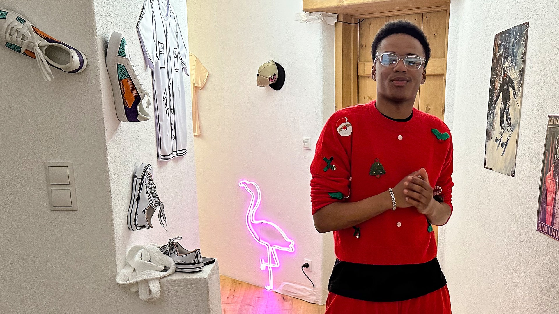 Ein Junge steht im Flur einer stylisch eingerichteten Wohnung. Er trägt einen roten Pullover und Brille. Der Flur ist mit Postern, einem pinkfarbenen Leucht-Flamingo und Sneakern dekoriert, die an der Wand hängen. Azad grinst cool in die Kamera.