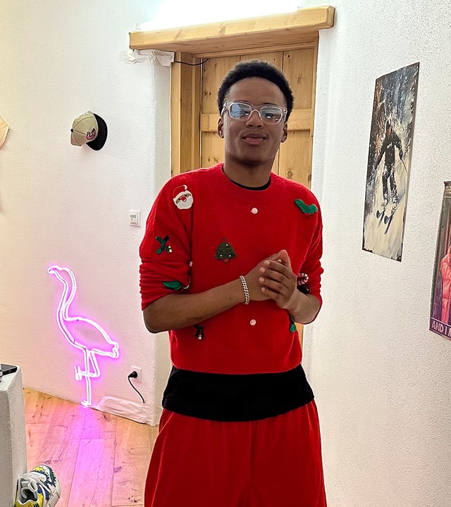 Ein Junge steht im Flur einer stylisch eingerichteten Wohnung. Er trägt einen roten Pullover und Brille. Der Flur ist mit Postern, einem pinkfarbenen Leucht-Flamingo und Sneakern dekoriert, die an der Wand hängen. Azad grinst cool in die Kamera.