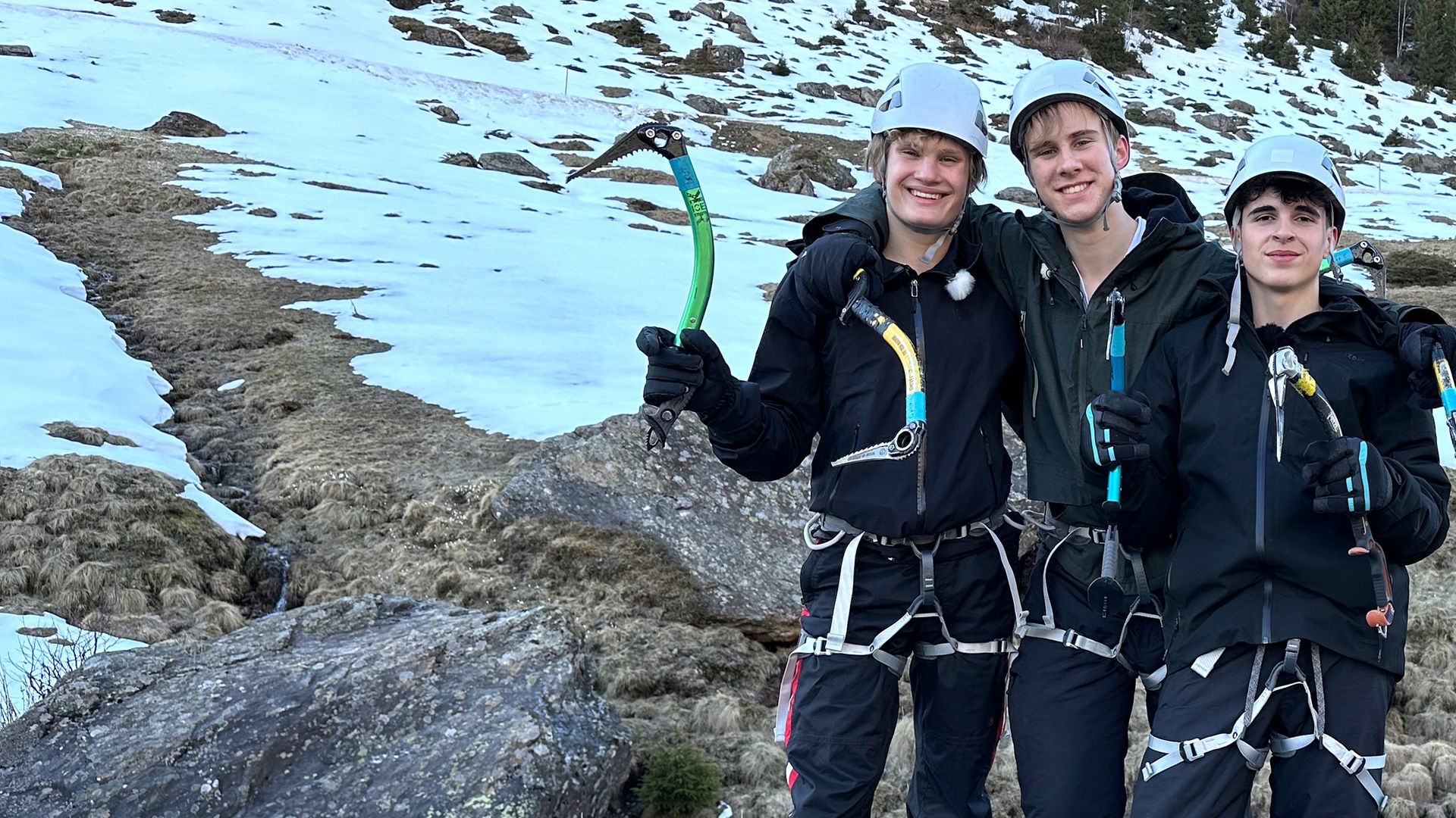 Drei Jungs tragen Klettergurte, Eispickel und Helme und stehen nebeneinander in einer felsigen und schneebedeckten Landschaft. Cay, Luke und Nick sind bereit fürs Eisklettern und grinsen in die Kamera.
