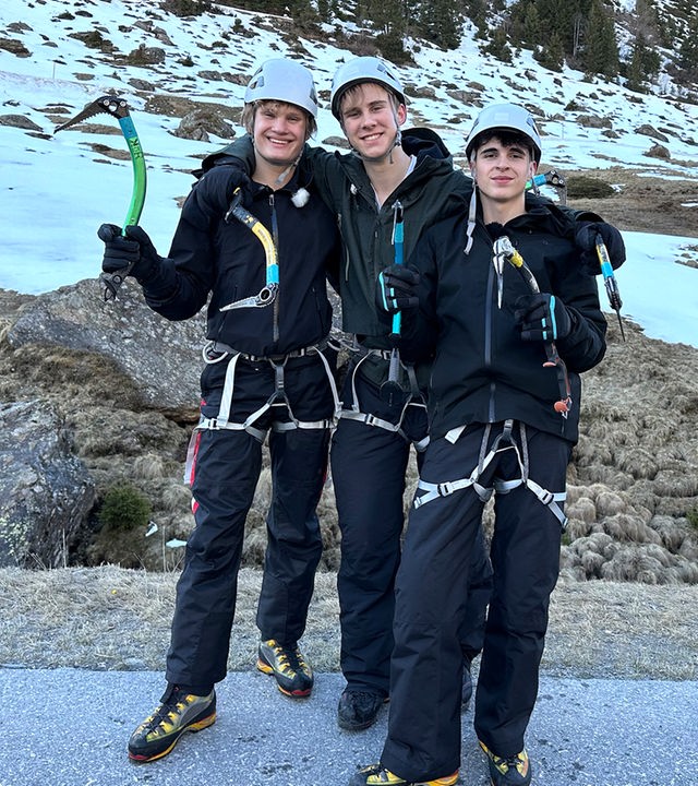 Drei Jungs tragen Klettergurte, Eispickel und Helme und stehen nebeneinander in einer felsigen und schneebedeckten Landschaft. Cay, Luke und Nick sind bereit fürs Eisklettern und grinsen in die Kamera.