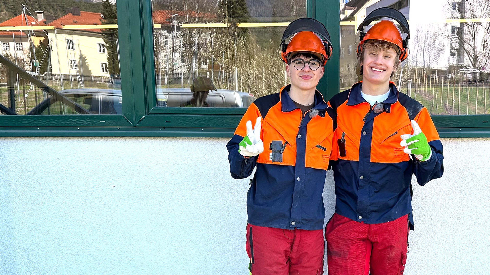Zwei Jungs in blau-weißen Overalls stehen nebeneinander und zeigen das Victoryzeichen. Es handelt sich um Sicherheitskleidung. Nick und Cay tragen zusätzlich orangefarbene Helme auf denen Schutz-Kopfhörer liegen und grün-weiße Arbeitshandschuhe.