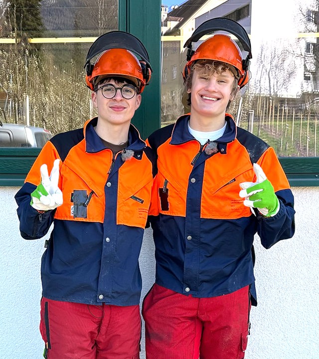 Zwei Jungs in blau-weißen Overalls stehen nebeneinander und zeigen das Victoryzeichen. Es handelt sich um Sicherheitskleidung. Nick und Cay tragen zusätzlich orangefarbene Helme auf denen Schutz-Kopfhörer liegen und grün-weiße Arbeitshandschuhe.