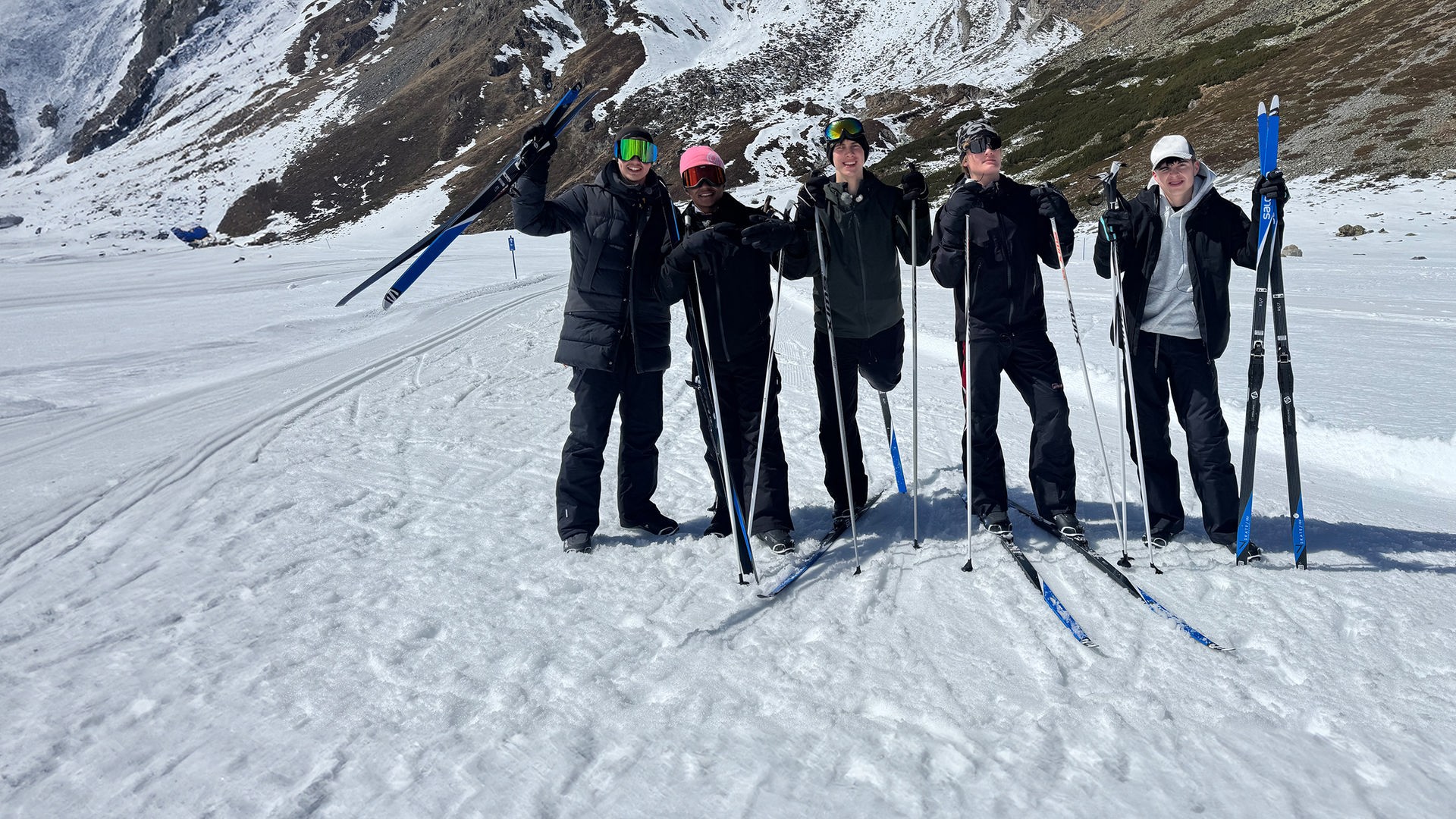 Fünf Jungs stehen in Skikleidung, mit Skibrillen und Mützen auf einer Skipiste. Sie tragen Langlauf-Skier an den Füßen oder halten sie in der Hand. Es sind Lorian, Azad, Luke, Cay und Nick aus der Jungs-WG 2026.