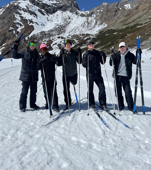 Fünf Jungs stehen in Skikleidung, mit Skibrillen und Mützen auf einer Skipiste. Sie tragen Langlauf-Skier an den Füßen oder halten sie in der Hand. Es sind Lorian, Azad, Luke, Cay und Nick aus der Jungs-WG 2026.
