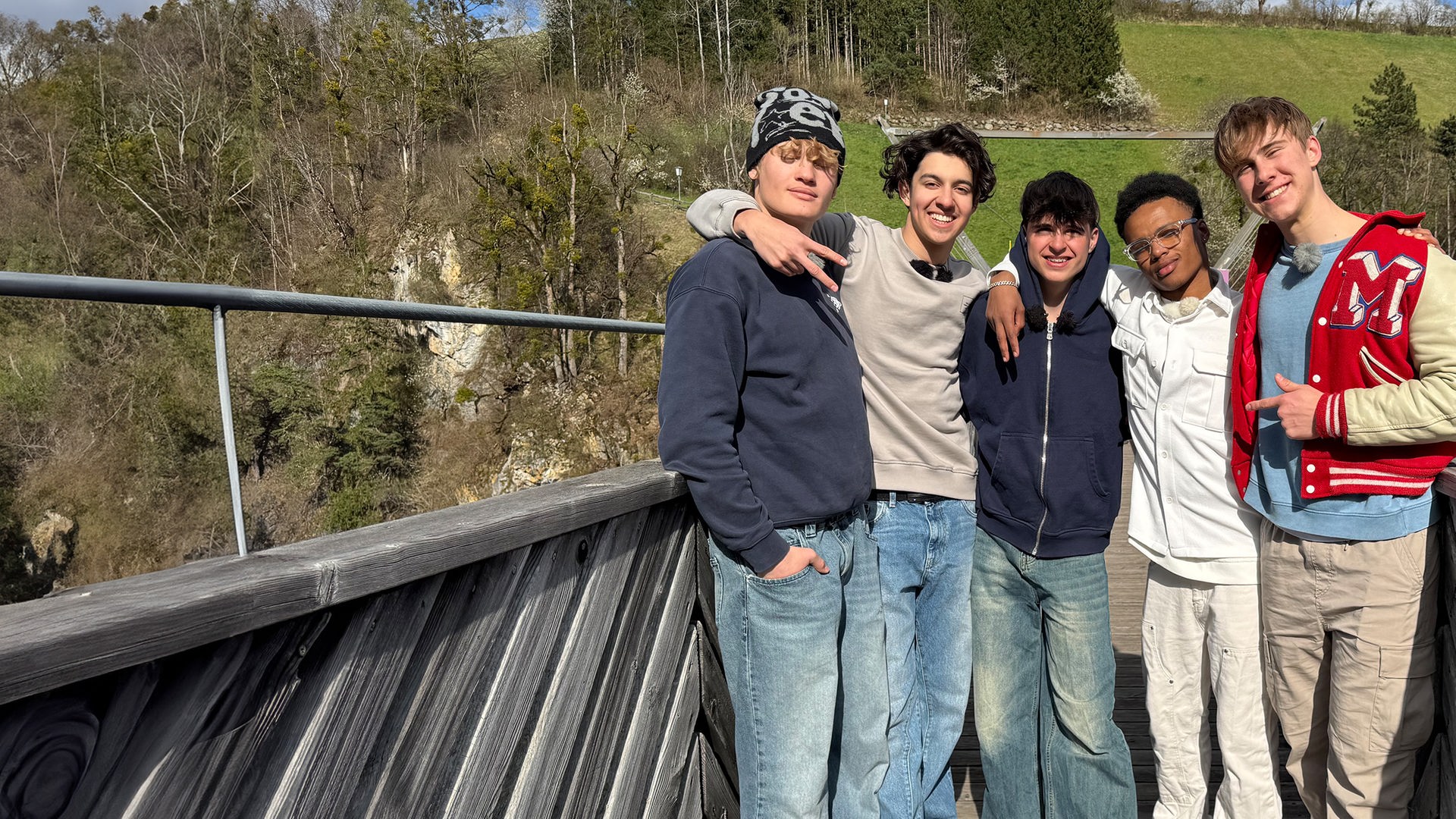Fünf Jungs stehen auf einer Holzbrücke nebeneinander. Im Hintergrund sind Tannen und grüne Wiesen zu sehen. Die Jungs tragen Jeans und Pullover oder Sweatshirt-Jacken. Sie haben die Arme umeinander gelegt und schauen fröhlich in die Kamera.